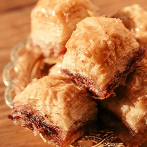 Close-up of golden, flaky baklava pieces with nut filling, glistening with syrup in a decorative bowl.