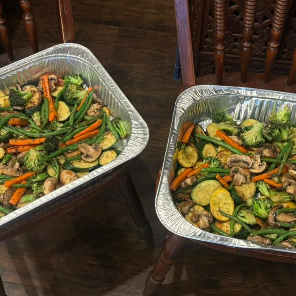 Two large foil pans filled with roasted vegetables, including broccoli, carrots, zucchini, and mushrooms.