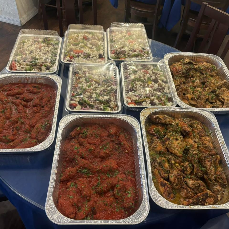 Multiple foil trays of Greek food, including salads with feta, chicken, and meatballs in red sauce.
